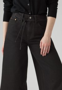 Sorte bredbenede jeans laget av denim, med beltestropp i midjen, subtile sømdetaljer og frontlommer.