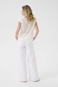 Blouse blanche en œillets avec des volants aux épaules, dos transparent, ourlet festonné, associée à un pantalon blanc fluide. Palette de couleurs neutres.