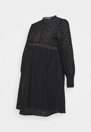 Vestido de maternidade preto em renda, com mangas longas, textura semi-translúcida, tecido com padrão em diamante e um decote alto com detalhes intrincados.