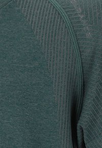 Grünes Sportshirt mit strukturierten Einsätzen und glatter Oberfläche, mit Kontrastnähten und einem Logo auf der Schulter.