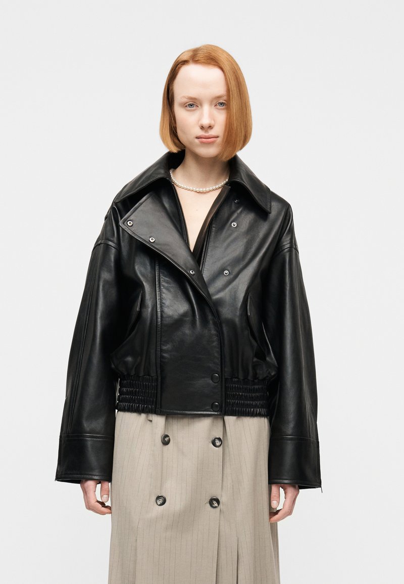 Νεαρή γυναίκα με κοντά κόκκινα μαλ λιά φοράει μαύρο δερμάτινο μπουφάν, μπεζ φούστα με λεπτές ρίγες και κολιέ με πέρλες σε απλό φόντο.