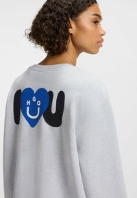 Grijze sweatshirt met een blauwe hartafbeelding en de tekst "I love U" met zwarte handontwerpen op de achterkant. Klassieke pasvorm met een zachte textuur.