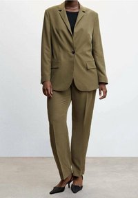 Abito su misura verde oliva con giacca aderente dotata di rever e tasche laterali, abbinato a pantaloni slim-fit e tacchi neri.