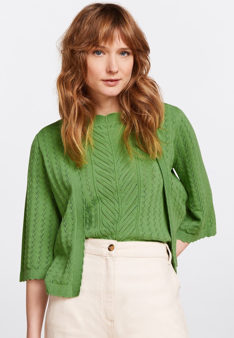 Suéter verde de punto con textura de patrón de hojas, con un cuello redondo y mangas hasta los codos, usado sobre una camiseta de color claro y pantalones.