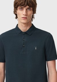 Dunkelblaues Poloshirt aus strukturiertem Stoff, mit einer Drei-Knopf-Leiste und einem kleinen gestickten Logo auf der linken Brust.