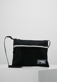 Borsa a tracolla nera in rete con zip bianca, cintura regolabile e patch blu del logo TOMMY JEANS sul davanti. Superficie testurizzata e design compatto.