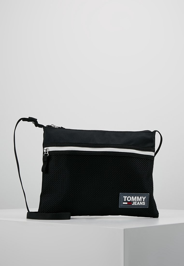 Borsa a tracolla nera in rete con zip bianca, cintura regolabile e patch blu del logo TOMMY JEANS sul davanti. Superficie testurizzata e design compatto.