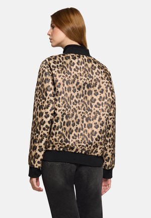 Vrouw die zijwaarts staat, gekleed in een jasje met luipaardprint met zwarte manchetten en kraag, gecombineerd met donkergrijze jeans tegen een effen achtergrond.