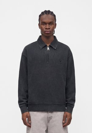 Giovane uomo con capelli intrecciati che indossa un pullover nero a maniche lunghe con zip a quarto e pantaloni grigio chiaro, in piedi davanti a uno sfondo bianco uniforme.