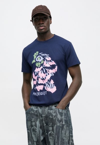 Μπλε βαμβακερό μπλουζάκι με πολύχρωμο γραφικό ενός πλάσματος σε ροζ, πράσινο και μπλε. Το μοντέλο το συνδυάζει με γκρι παντελόνι με σχέδια.