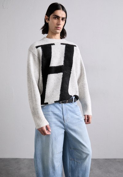 Γκρι πλεκτό πουλόβερ με μεγάλο μαύρο σχέδιο "Η", ριμπ μανσέτες, συνδυασμένο με ανοιχτό μπλε παντελόνι τζιν σε φαρδύ κόψιμο με ραμμένα αξεσουάρ.