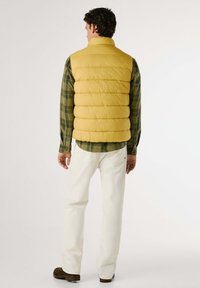 Gilet imbottito giallo con alto colletto, indossato sopra una camicia a quadri verde. Pantaloni bianchi e scarpe marroni completano l'outfit. Vista posteriore.