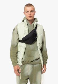 Outfit verde e nero con un gilet puffo verde chiaro, felpa con cappuccio, pantaloni abbinati e una borsa a tracolla nera indossata sul petto.