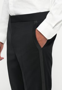 Schwarze maßgeschneiderte Hose mit einer eleganten, flachen Vorderseite und einer Seitentasche. Sie zeichnet sich durch eine glatte Textur und dezente Satinakzente entlang der Taschenkante aus.