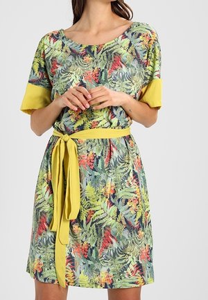 Frau trägt knielanges Kleid mit Blumenmuster, gelbem Gürtel und hochgekrempelten kurzen Ärmeln, steht mit gefalteten Händen auf der Taille.