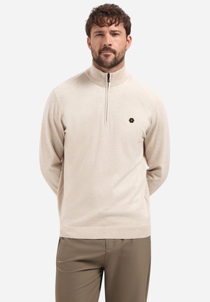 Pull beige zippé avec un col montant, en tissu lisse, présentant un petit logo hexagonal sur la poitrine et des poignets côtelés.