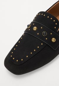 Mocassino in suede nero con punta rotonda, caratterizzato da borchie decorative oro e nere lungo la parte superiore e una cintura in pelle liscia.