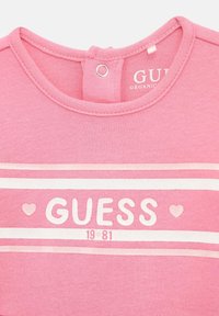 T-shirt en coton rose avec un col rond, des accents rayés blancs et le logo "GUESS" avec des cœurs en rose pâle sous le col.