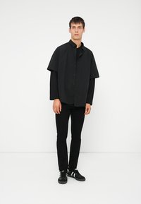 Tânăr îmbrăcat cu un tricou cu mâneci lungi, în straturi, cu un overlay cu mâneci scurte, pantaloni negri și adidași negri cu dungi albe.
