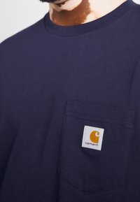 Camiseta de algodón azul marino con escote redondo y un bolsillo en el pecho izquierdo que presenta una etiqueta del logo de Carhartt en blanco y naranja.