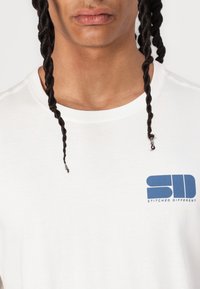 Vit bomull T-shirt med en blå logotyp "SD" och "STITCHED DIFFERENT" på vänster bröstområde; kort ärm och rundad halsdesign.