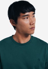 Dunkelgrünes Baumwoll-T-Shirt mit Rundhalsausschnitt und kurzen Ärmeln. Verfügt über ein kleines, auf der linken Brust gesticktes Logo. Glatte Textur.