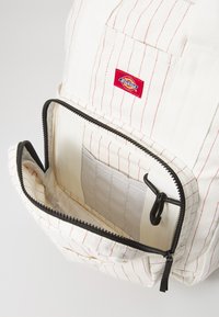 Rucsac din material alb cu dungi roșii, având un buzunar frontal cu fermoar și fermoar negru, plus o etichetă din material cosută pe față.