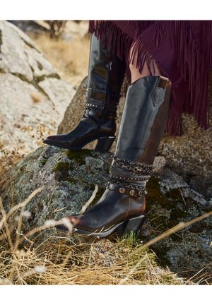Des bottes en cuir noir montant jusqu'au genou, avec un bout pointu et un talon épais, ornées d'accents métalliques décoratifs et de sangles texturées.