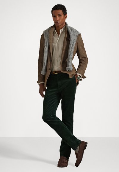 Blazer en vichy marron sur une chemise blanche à boutons crème, pantalon en velours côtelé vert, pull en tricot gris, et mocassins marron. Montre en argent au poignet.