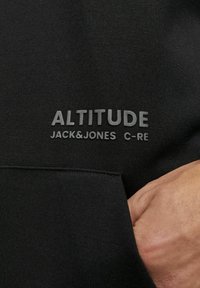 Sort sweatshirt med en frontlomme, struktureret stof og hævet "ALTITUDE JACK&JONES C-RE" branding i en kontrastfarve.