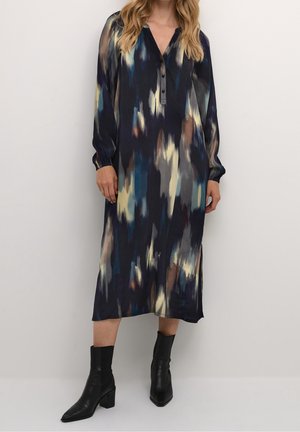 Femme portant une robe midi à manches longues avec un motif abstrait multicolore foncé et des bottines noires, debout devant un fond uni.