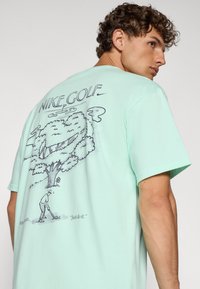 Lys grøn bomulds T-shirt med en grafisk illustration af en golfspiller og et træ på bagsiden, med "NIKE GOLF" ovenfor i fed skrift.