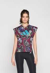 Haut à manches courtes avec motif graphique de papillon en violet et noir ; accent turquoise sur la poitrine ; fabriqué à partir d'un tissu léger et extensible.