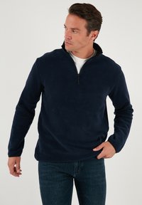 Pullover in fleece blu navy con colletto alto e design a mezza zip. Texture morbida e maniche lunghe. Abbinato a jeans in denim scuro.