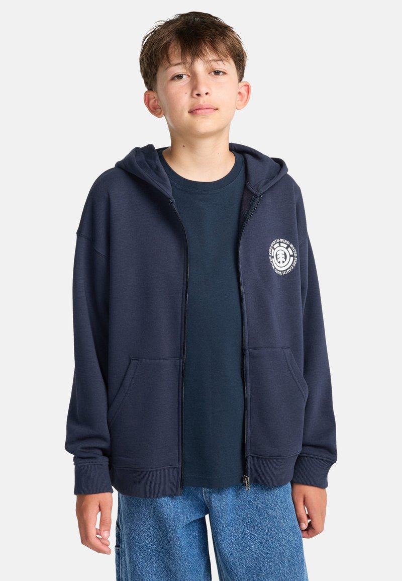 Tmavomodrá mikina na zips s kapucňou na šnúrku, prednými vreckami a bielym logom na ľavej strane hrudníka; nosená cez tmavomodrú košeľu a modré džínsy.