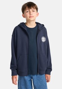 Sudadera con cremallera azul marino, hecha de una mezcla de algodón, que cuenta con un bolsillo canguro, puños acanalados y una impresión de logo en el lado izquierdo del pecho. Combinada con una camiseta azul marino.