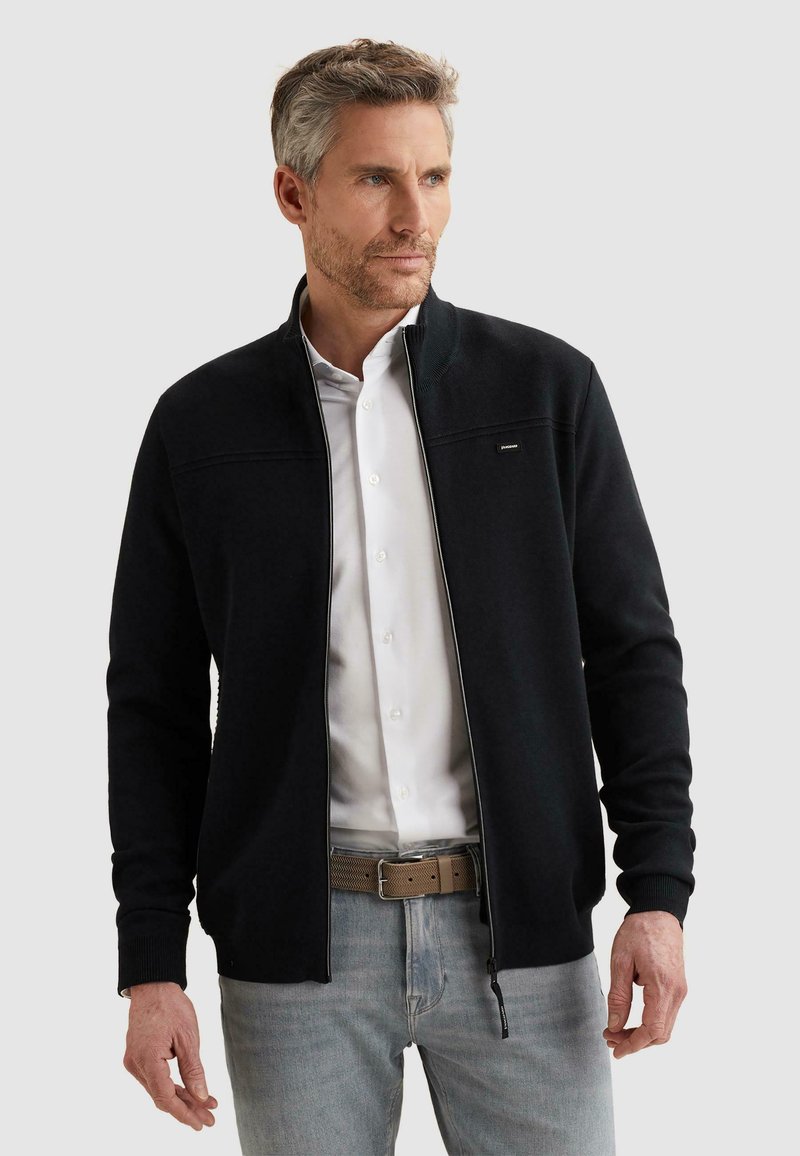 Homme d'âge moyen portant une veste noire à fermeture éclair par-dessus une chemise blanche et un jean gris clair, debout devant un fond uni.