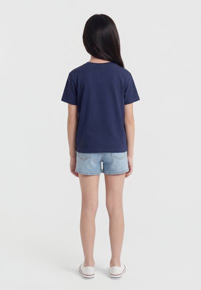 Barn med langt mørkt hår iført en marineblå t-skjorte, lyse denimshorts og hvite joggesko som står med ryggen mot på hvit bakgrunn.