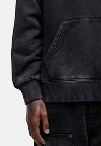 Hoodie in denim nero con tasca a marsupio sul davanti, polsini e orlo a coste. Il tessuto testurizzato presenta una leggera sbiaditura. Mano con tatuaggi visibili.