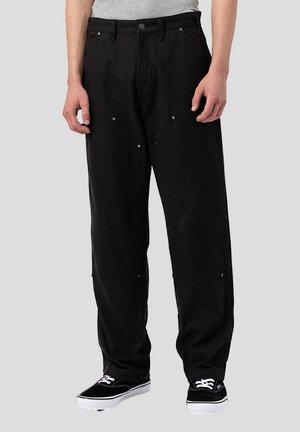 Homme portant un pantalon noir à jambes larges avec plusieurs poches et des baskets noires à lacets blancs sur fond gris clair.