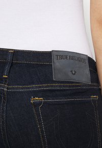 Jeans de mezclilla azul oscuro con costuras amarillas; presentan un parche de cuero gris con el logo "TRUE RELIGION" en la parte trasera.