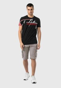 Black t-shirt with white and red graphic print, paired with gray striped cargo shorts. Model wears white sneakers, standing on a neutral background.