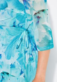 En blå blommig omslagsklänning med genomskinliga ärmar, som har ett knytband i midjan och en mjuk textur. Tyget har ett livfullt, abstrakt blommönster.