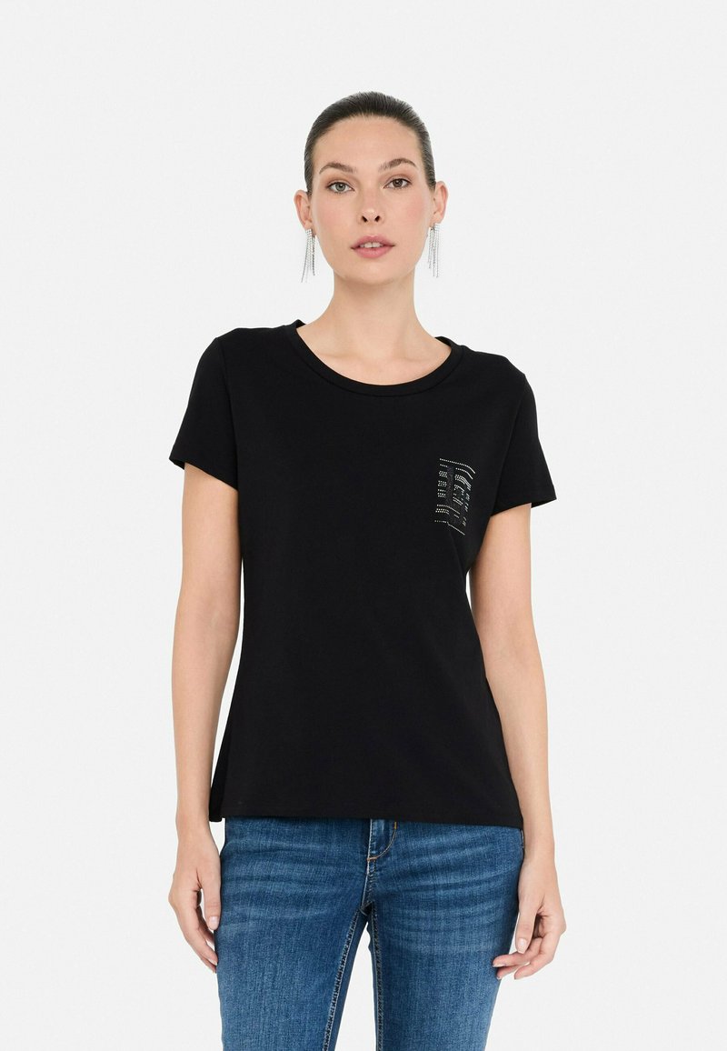Kvinde iført en sort T-shirt med korte ærmer og et lille rhinestones mønster på brystet samt blå jeans, stående foran en hvid baggrund.