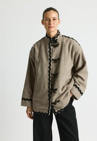 Brown herringbone fabric jacket with black scalloped trim, lax fit, high collar, and traditional toggle fastenings. Features wide sleeves.