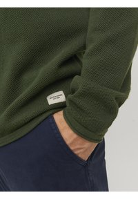 Mörkgrön strukturerad tröja med ribbade ärmslut och en etikett med texten "JACK & JONES EST. 1990" vid fållen, tillsammans med marinblå byxor.