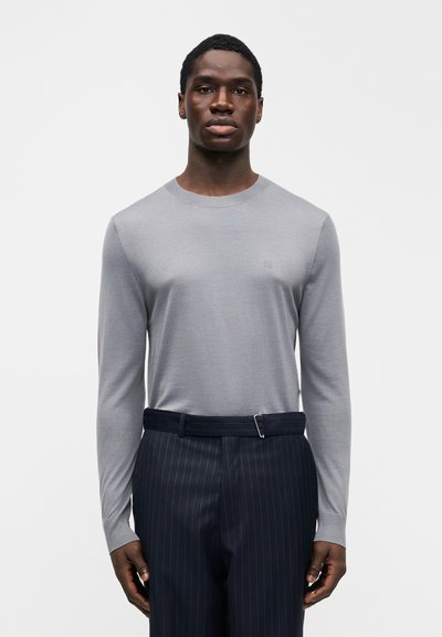 Haut gris à manches longues en tissu doux, avec un col arrondi et un logo discret, associé à un pantalon bleu marine à fines rayures.