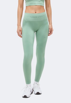 Femme portant des leggings Nike Pro vert clair et un crop top assorti, associés à des baskets Nike blanches et des chaussettes blanches, debout sur fond blanc.
