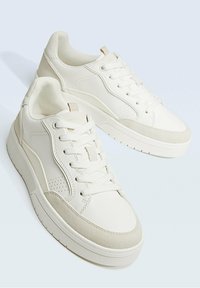 Un paio di sneakers bianche basse con design a lacci, suola testurizzata e accenti in camoscio su uno sfondo chiaro.