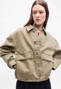 Γυναίκα που φοράει φαρδιά μπεζ ζακέτα με κουμπιά και μεγάλες τσέπες με καπάκι, τα χέρια στις τσέπες, κοιτάζοντας προς τα δεξιά σε λευκό φόντο.